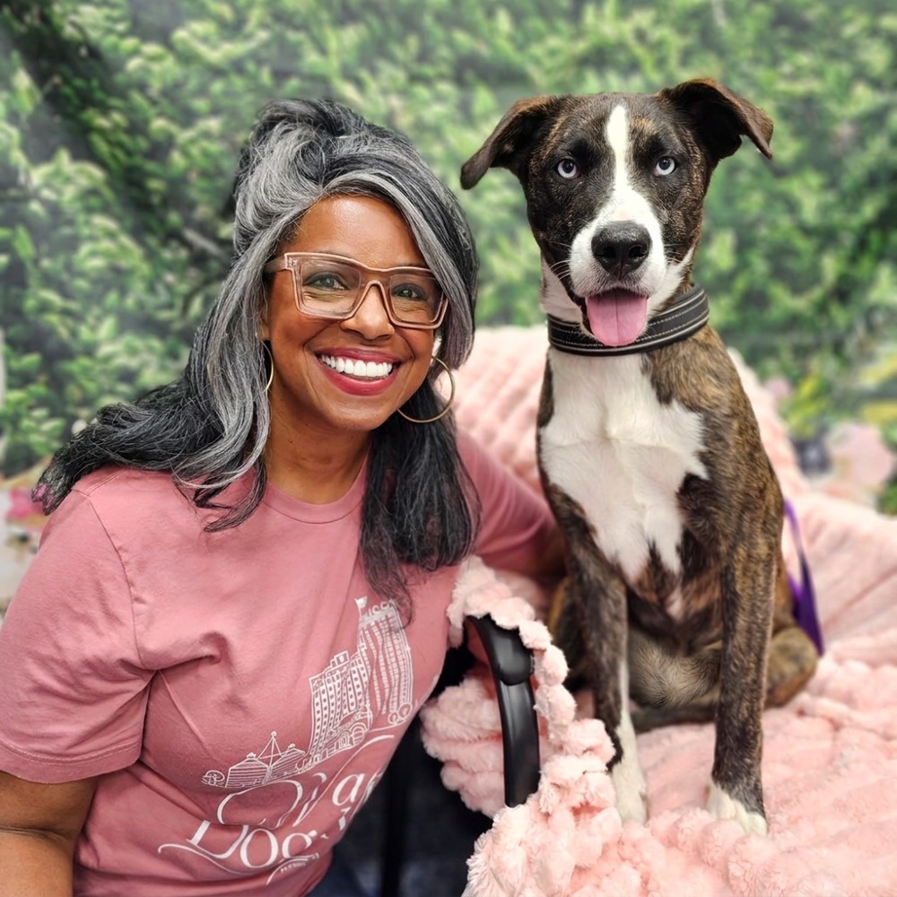 Jackye Clayton smiling beside her dog Shiloh, a brindle dog with pale blue eyes and white markings
